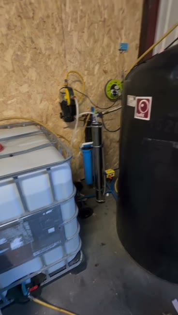 KBG's pure water system in its yard: the pump, filter housing and reverse osmosis unit on the wall, the TDS meter above them, an IBC of waste water on the left and the black pure water storage tank on the right